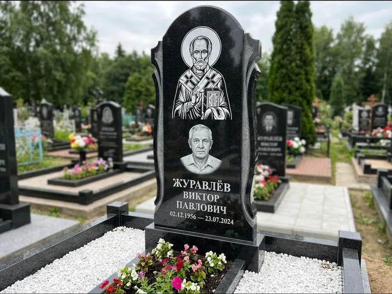 Памятник с гравировкой иконы: стела с образом в полный рост на обороте — гравировка образа Матроны Московской, низкие кусты спиреи в цвету, работа «Лебедева» в Белгороде
